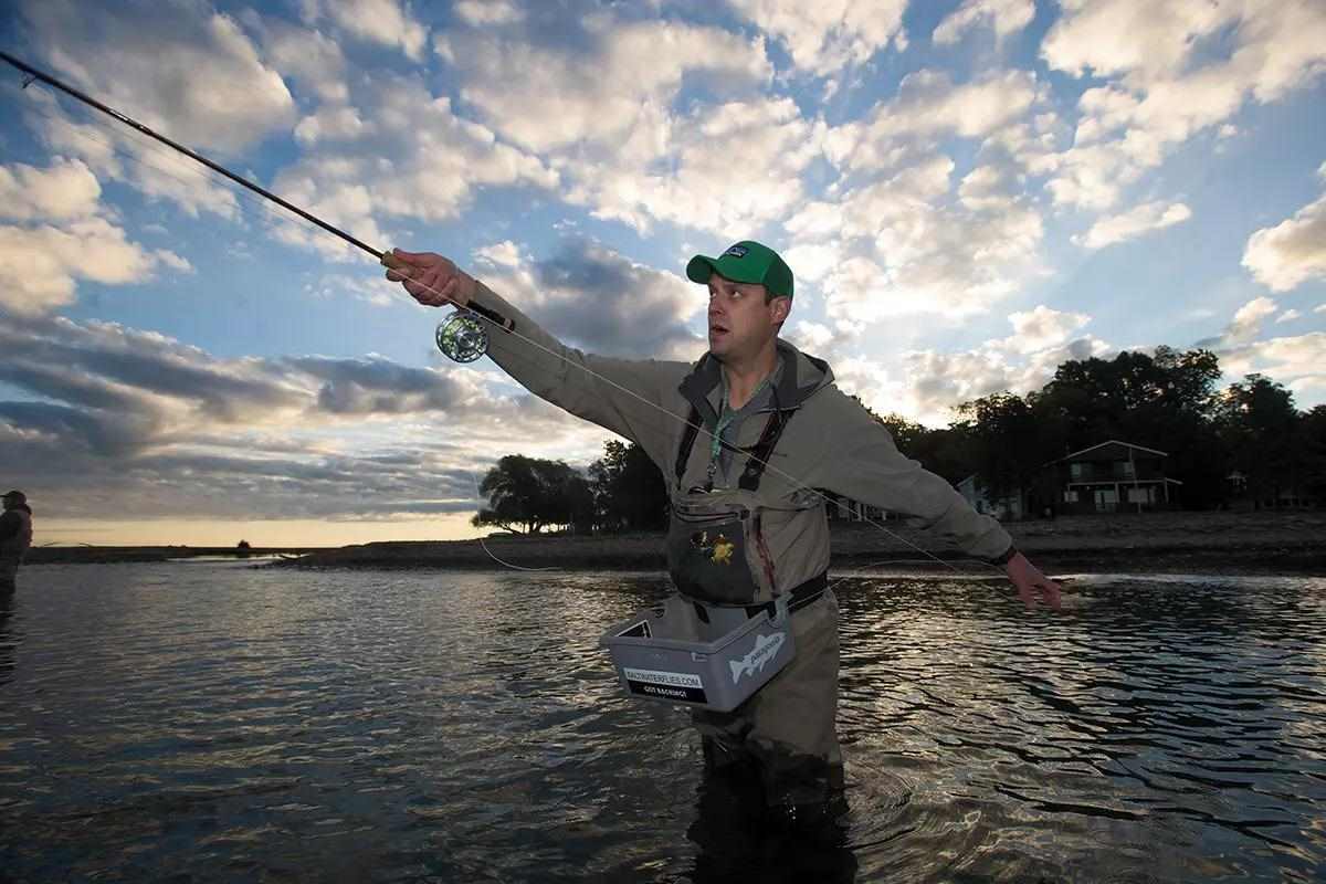 A fly angler casting in the evening light, watching his rod tip, wearing a stripping basket. 