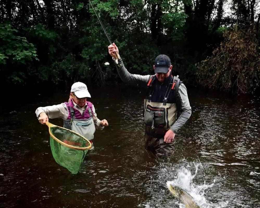 A fly angler battling a jumping fish with an angler standing by with a landing net.