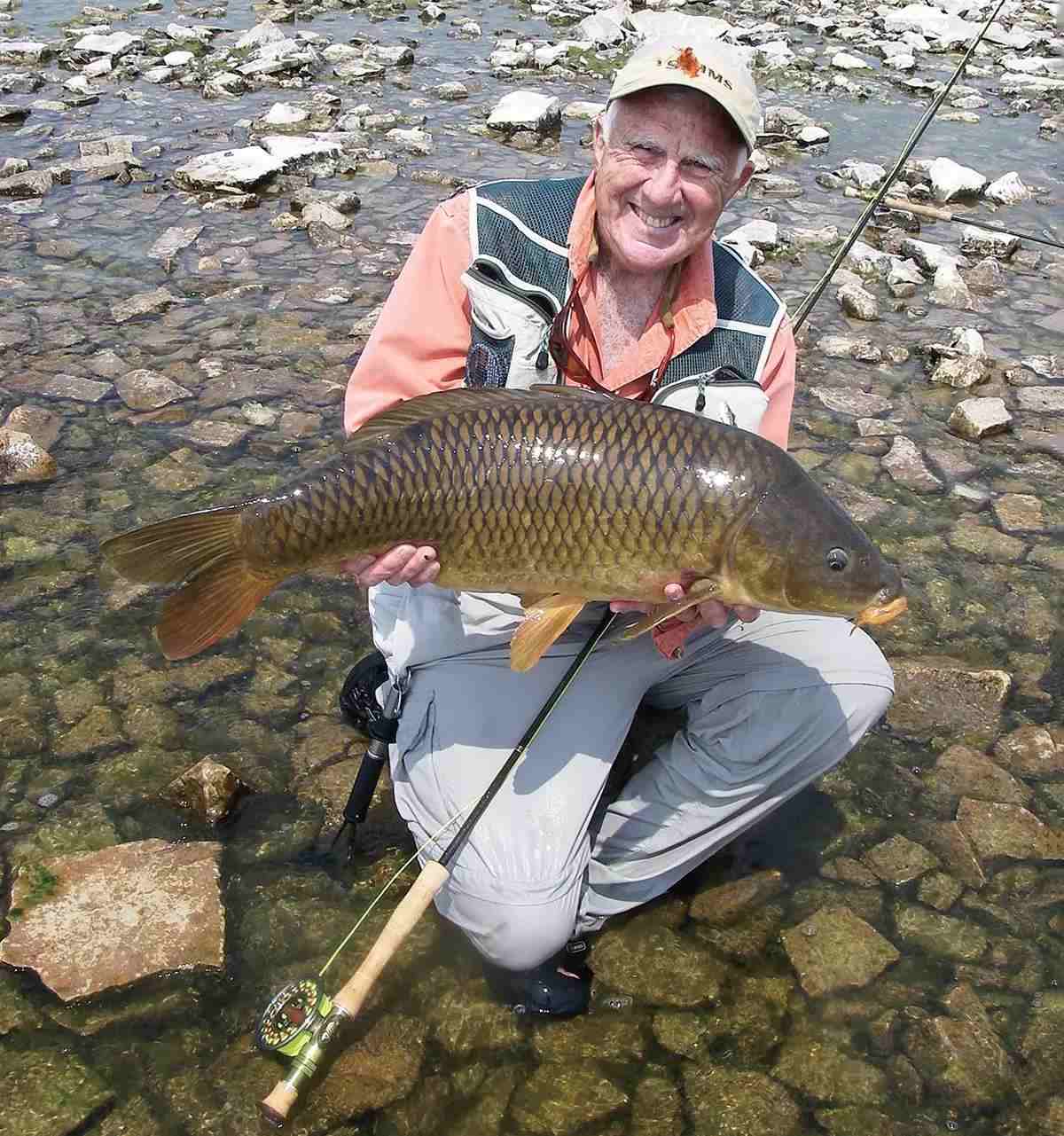 Dave Whitlock smiling, kneeling, and holding a large carp. 