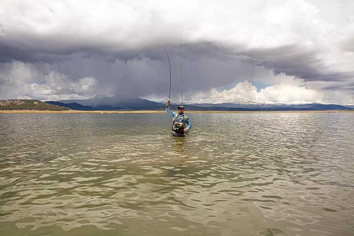 A fly angler wading waist deep in a lake holding his rod high fighting a fish under stormy skies.