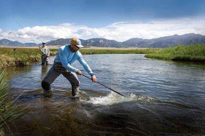 A man netting a trout for a fly angler in a small stream in a mountainous landscape.
