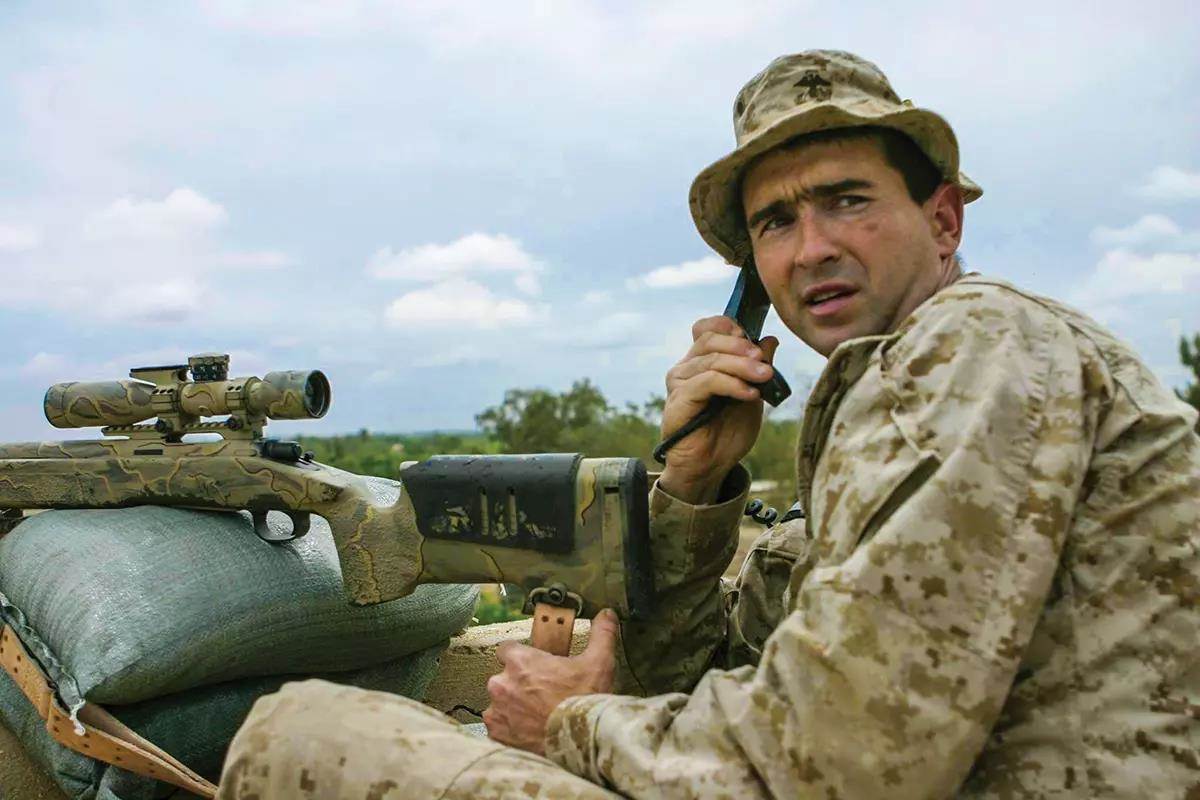 A marine sniper in desert camo on a phone call next to his set-up rifle. 