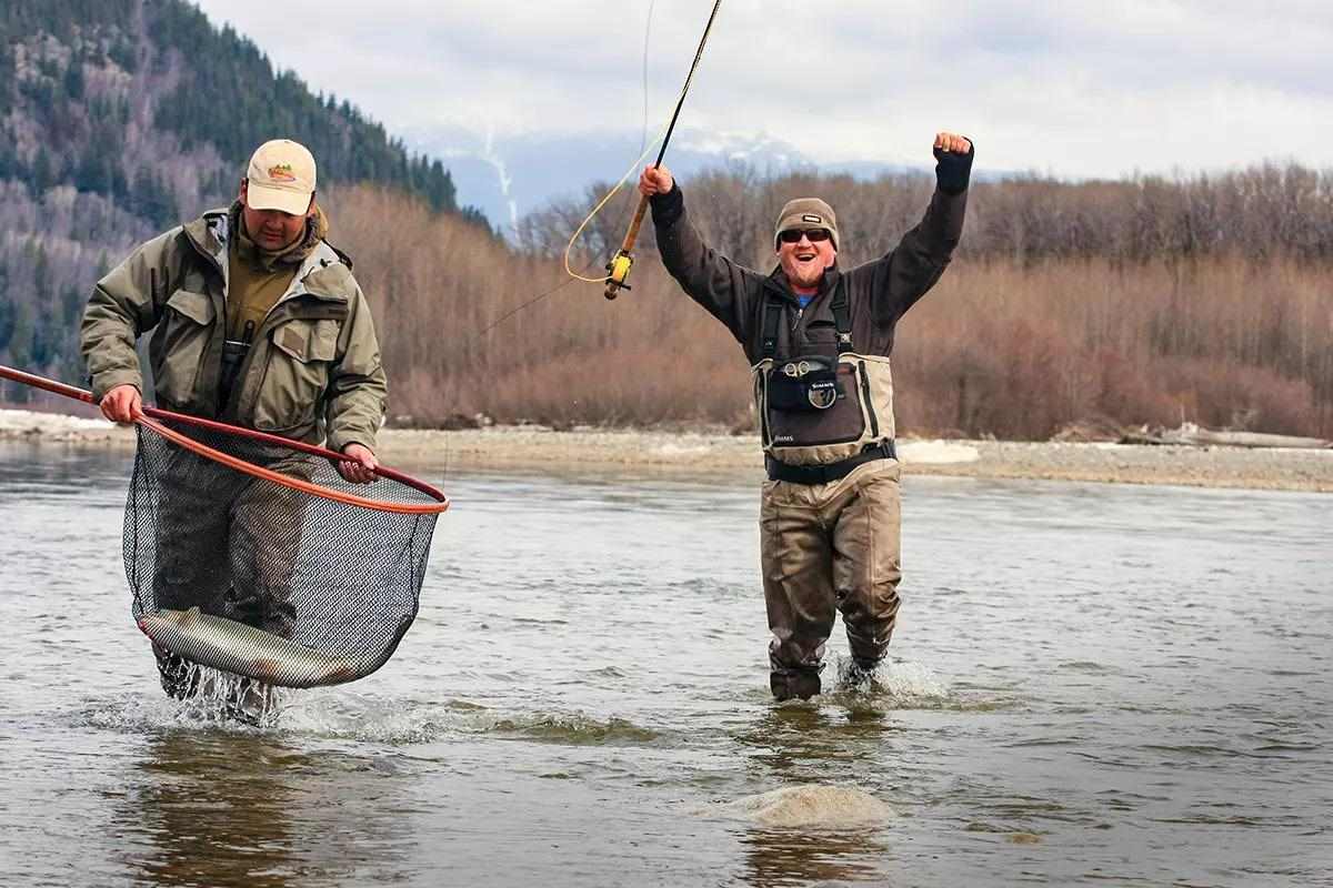 Two fly anglers wading ankle deep in a river, one holding a net with a big steelhead, the other celebrating. 
