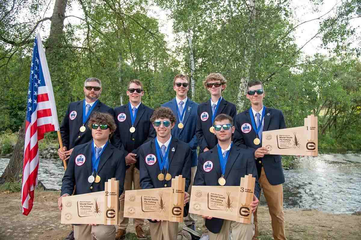 Eight young men in blue sport coats holding trophies and an American flag in front of a river.
