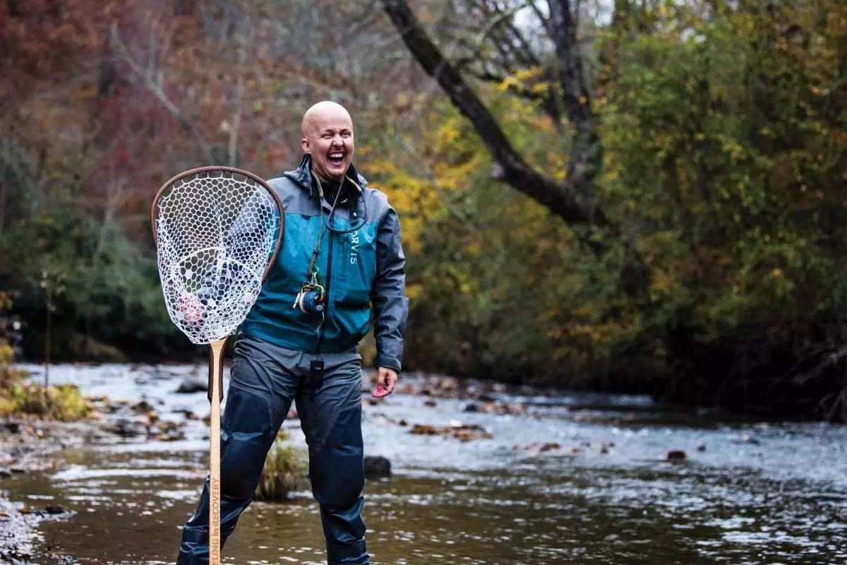 A bald woman laughing, wearing waders and holding a boat net on a river.