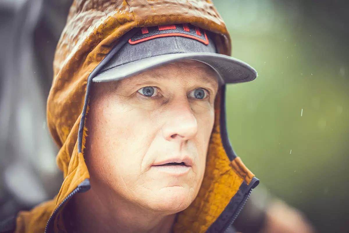 A close-up of the face of man with an orange hood and blue cap on. 