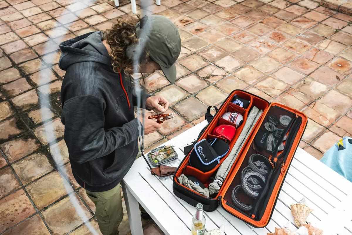 A man ties a fly onto his line standing on square tiles next to a white table with his gear on it. 