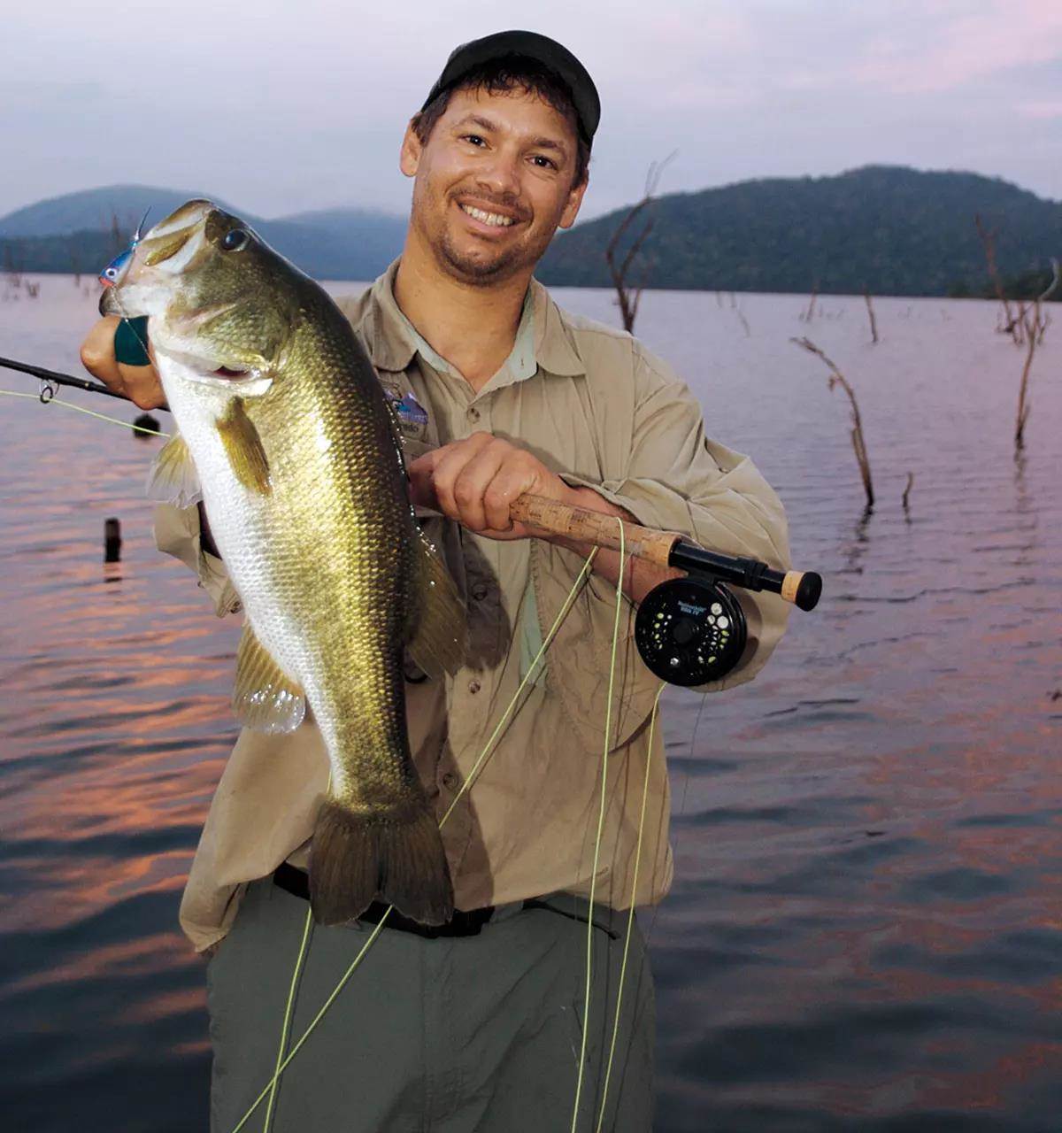 A smiling man on a lake with dead trees holding a fly rod and a large largemouth bass.