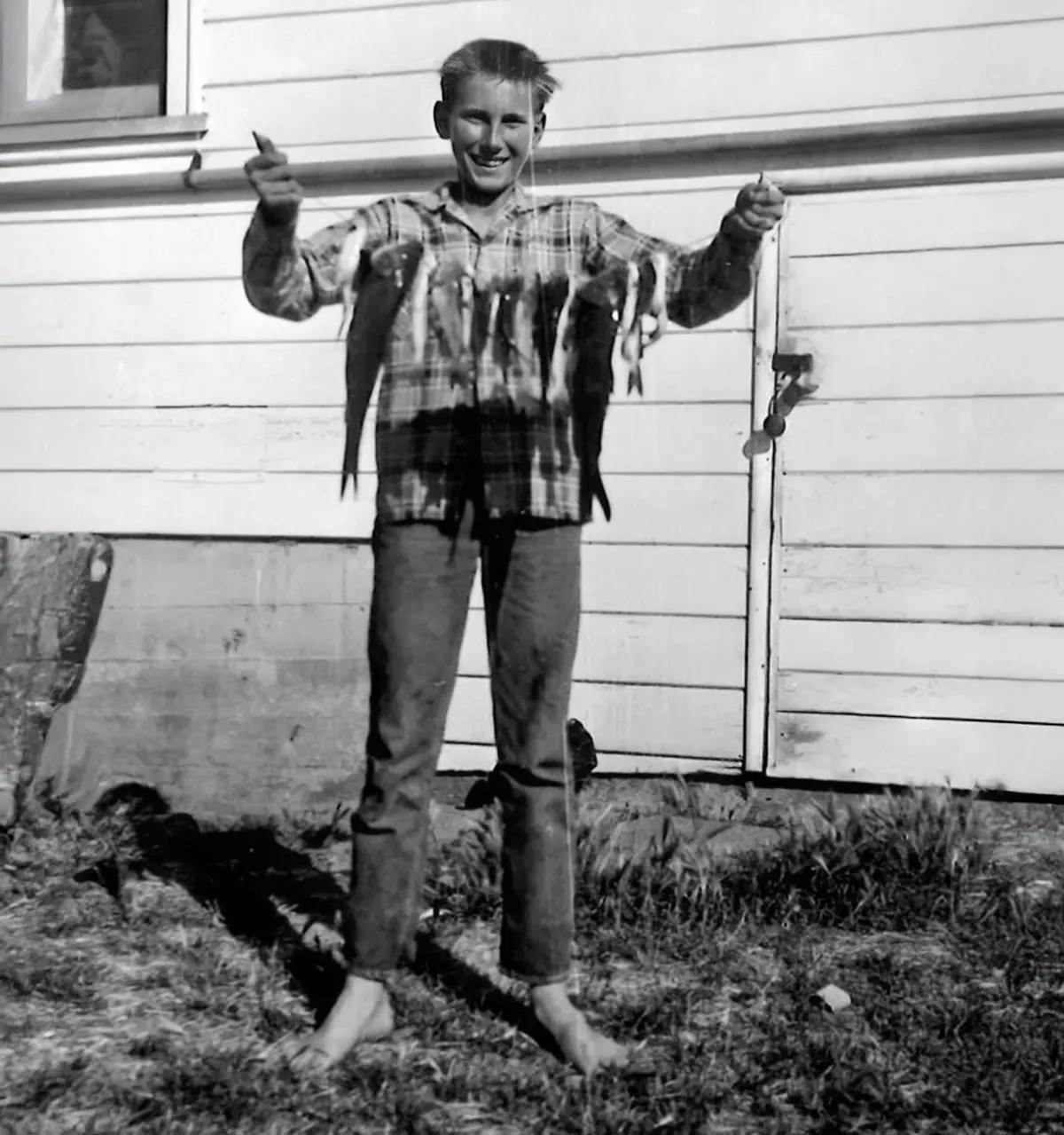 An old-timey black-and-white image of a young man holding a stringer of fish in front of a white house.