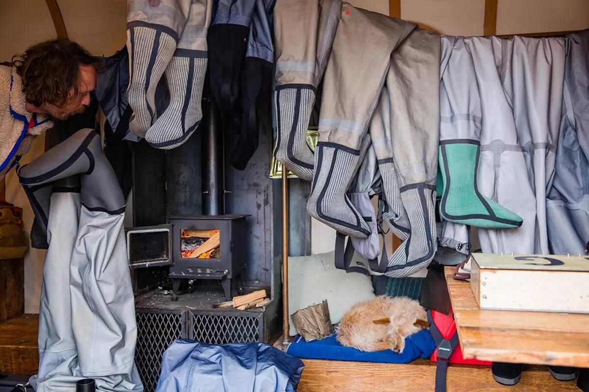 A man peeking in from the left holding a pair of inside-out waders next to a mini wood-burning stove; a clothesline full of inside-out waders is to the right. 