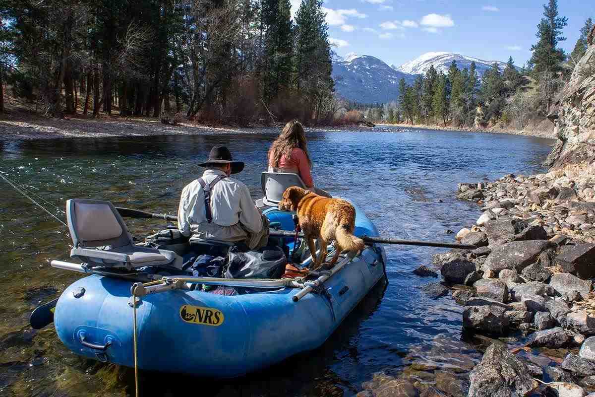 Two anglers in a blue river raft on a river in the mountains, rigging up. A brown dog named Boges stands on the gunwale.