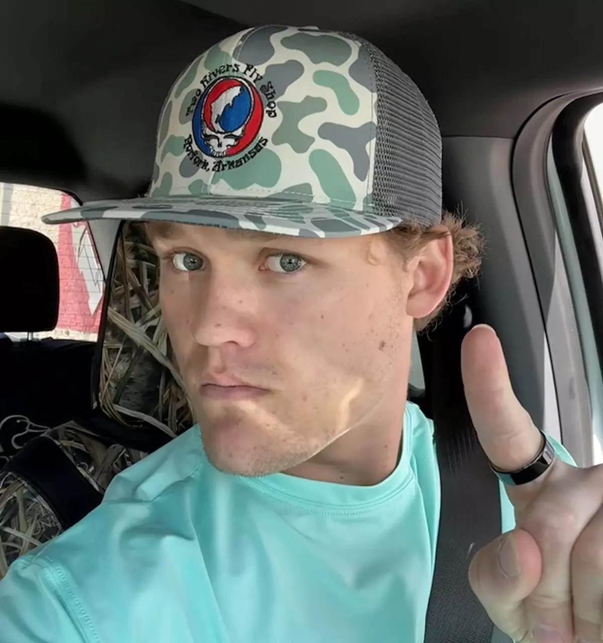 A young man in the driver's seat of a vehicle pointing up at his camo baseball cap.