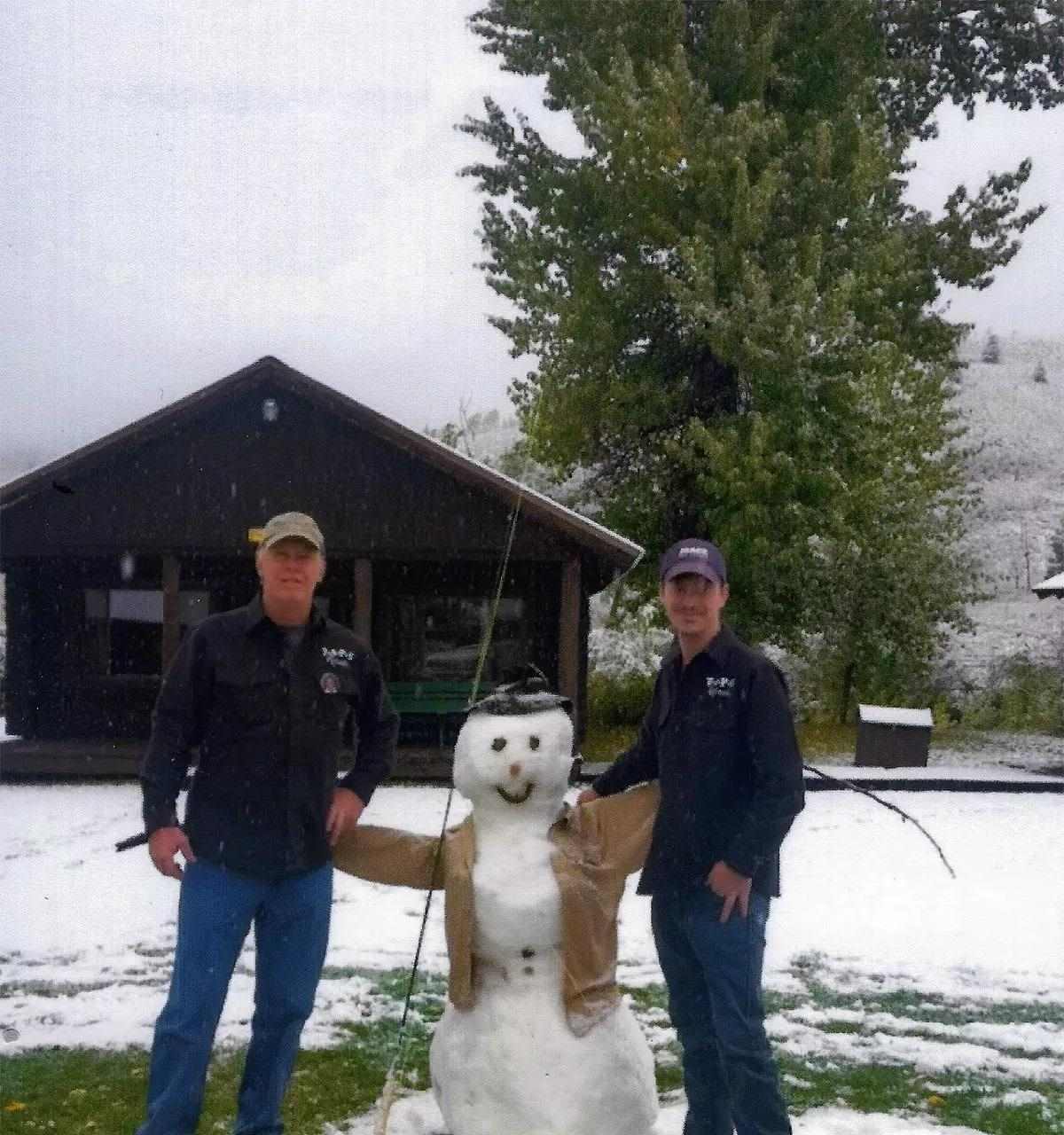 Two adult men posing with a snowman dressed up as a fly angler in front of a cabin.