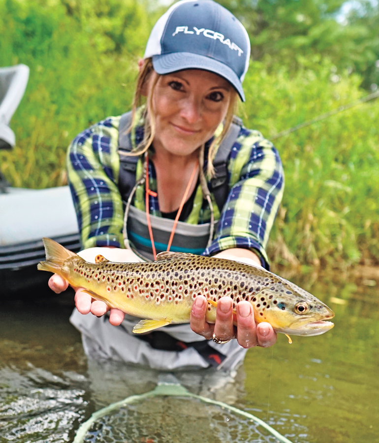 woman wearing flannel shirt and waders looking at camera holding brown trout near camera